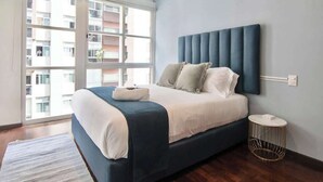 Een moderne slaapkamer met een groot bed, een rond bijzettafeltje en uitzicht op een gebouw door een groot raam.