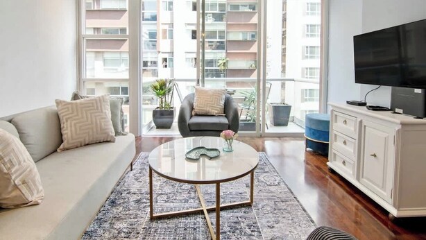 Een moderne woonkamer met een bank, een ronde salontafel, een televisie en uitzicht op een balkon met planten.