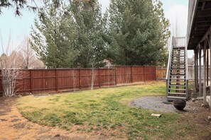 Backyard features a fire pit, ladder, and wooden fence, surrounded by trees and lush greenery.