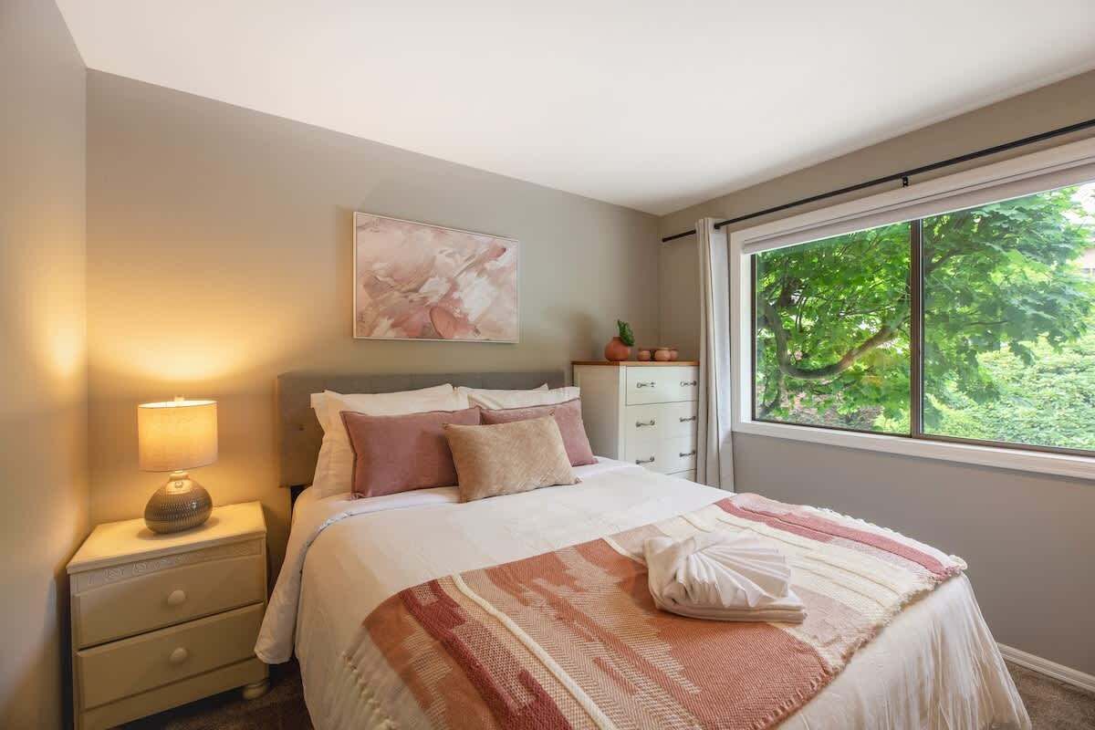 A bedroom with a bed, bedside table, lamp, and a window with a view of greenery.