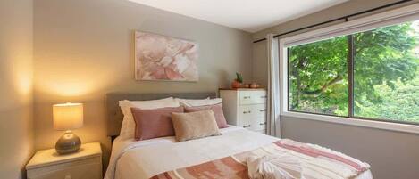 A bedroom with a bed, bedside table, lamp, and a window with a view of greenery.