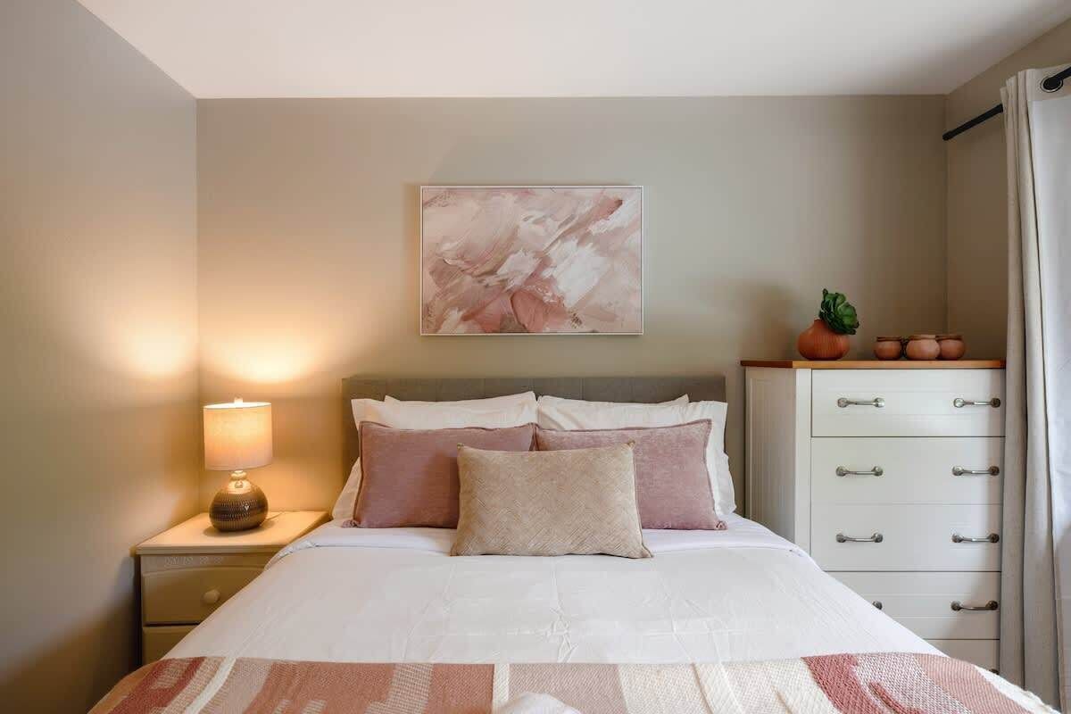 Una camera da letto ordinata con un letto, una lampada da comodino, una cassettiera e un quadro astratto appeso alla parete.