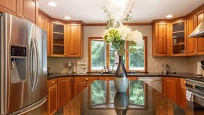 A modern kitchen with wooden cabinets, a stainless steel refrigerator, a large island with a vase of flowers, and a window providing natural light. - Serene 3b2b Home, Family-Friendly, Backyard & A/C (Redmond)