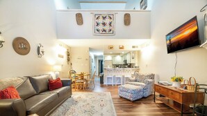 Amplia sala de estar con sofá, reposapiés y una mesa de centro. Se observa una cocina al fondo con armarios blancos y una zona de comedor.