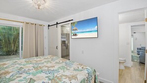 Une chambre à coucher avec un lit, un tableau représentant une plage, une porte donnant sur une salle de bain et une vue sur des palmiers à l’extérieur.