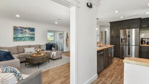 A modern living room with a sofa, coffee table, and artwork. Adjacent is a kitchen with stainless steel appliances and wooden cabinetry. - 🌊🏖️ Coral Cove B | Screened Porch + Pool | Steps to Beach + Times Square (Fort Myers Beach)