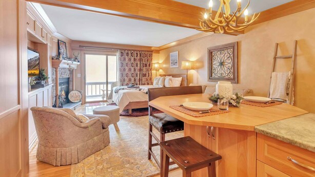 A cozy room with a wooden ceiling, a chandelier, a dining table with a bench, a fireplace, and a large window with curtains. - Cozy Studio with Fireplace, King Bed, A/C (Ellicottville)