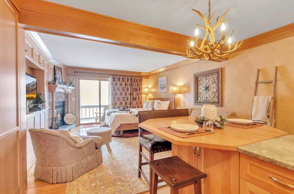 Una habitación acogedora con techo de madera, un candelabro, una mesa de comedor con un banco, una chimenea y un ventanal con cortinas.