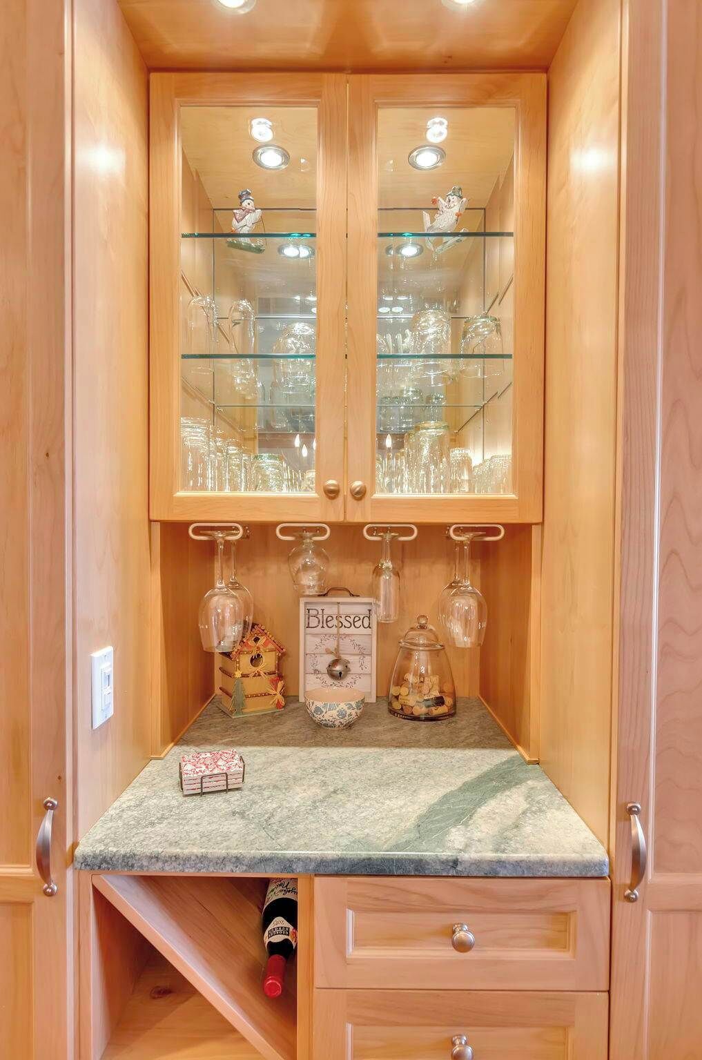 Ein Holzkabinett mit Glastüren, in dem Gläser, eine kleine Holzkiste, ein Schild mit der Aufschrift 'Blessed' und eine Flasche in einer Schublade aufbewahrt werden.