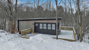 A small, single-story building with a dark door and windows, surrounded by snow-covered ground and trees.