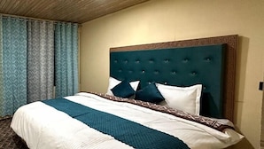 A neatly made bed with a patterned bedspread and pillows, a wooden headboard with a decorative border, and a blue curtain on the window. - Oasis Retreat Cottages Pangong (Leh)