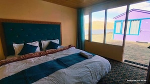 A bedroom with a large bed, a window with a view of a building, and a patterned bedspread. - Oasis Retreat Cottages Pangong (Leh)