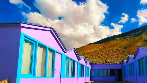 A row of purple and blue buildings with large windows, set against a mountainous backdrop. - Oasis Retreat Cottages Pangong (Leh)