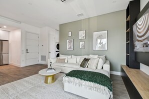 A modern living room with a white sofa, a round coffee table, and a green throw blanket. There are framed pictures on the wall and a built-in shelf with a mirror.