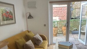 Un salon avec un canapé de couleur moutarde, une petite table et une porte-fenêtre coulissante donnant sur un espace extérieur agrémenté d’un mur en briques et de plantes en pot.