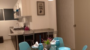 A modern kitchen with a dining area. It includes a kitchen island with a sink, a stove, and cabinets. There is a framed sign on the wall that reads "It's Wine Time." The dining area has a glass table with blue chairs and a vase with flowers. - Beach, your apartment near the sea and the Mayan Train (Playa del Carmen)