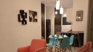 A modern living room with a red armchair, a glass dining table with blue chairs, and a kitchen area in the background. - Beach, your apartment near the sea and the Mayan Train (Playa del Carmen)
