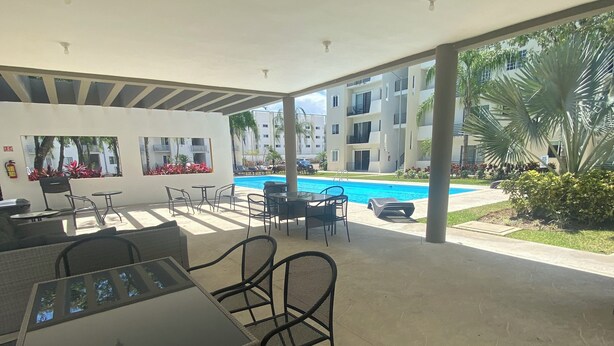 A covered outdoor area with tables and chairs, a pool, and a view of a residential building. - Beach, your apartment near the sea and the Mayan Train (Playa del Carmen)
