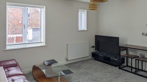 Uma sala de estar moderna com uma mesa de centro de vidro, sofá de couro, televisão e duas janelas com vista para um edifício de tijolos.