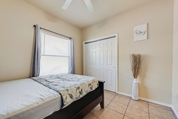 Quarto com cama, janela com cortinas, porta, placa na parede e um vaso com plantas secas.