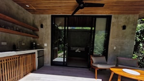 A modern kitchen with a built-in oven, a coffee machine, and a sink. There is a wooden table and a chair outside, and a sliding glass door leading to a balcony with a sofa. - Serenity Wave Bungalow (Cahuita)