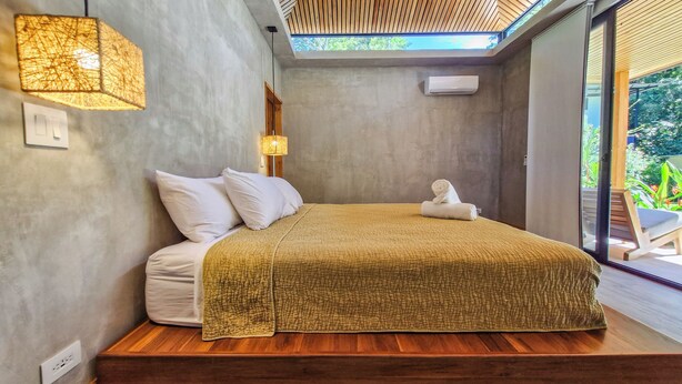 A modern bedroom with a wooden ceiling, a large bed with a textured blanket, a bedside lamp, and a view of the outdoors through a sliding door. - Serenity Wave Bungalow (Cahuita)