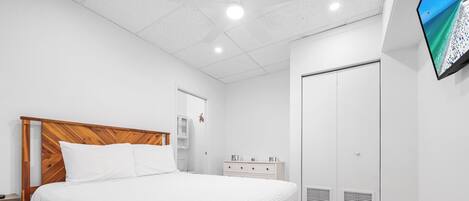 Un dormitorio con cama de madera, ropa de cama blanca, un armario blanco y un televisor en la pared.