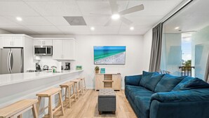 A modern kitchen with white cabinets, a stainless steel refrigerator, and a microwave. There is a wooden bench seating area with a grey ottoman in the center. A blue sofa is against the wall, and a large window with curtains is visible. - Perfect Cozy Island Get-Away on Siesta Key! (Siesta Key)