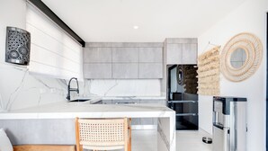 A modern kitchen with a marble island, stainless steel appliances, and a seating area with a wooden chair. - Romantic Getaway with Stunning Panoramic Ocean Views, Amazing Sunsets (Playa Flamingo)