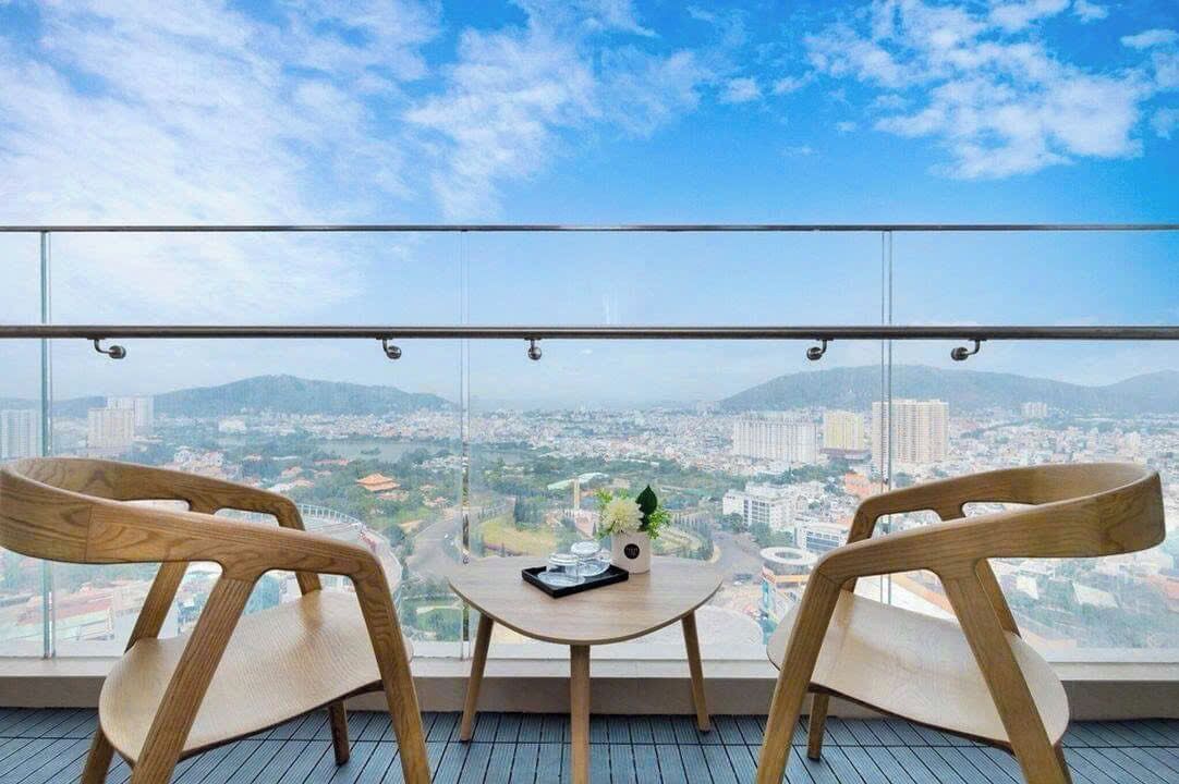 A balcony with two wooden chairs and a small table, overlooking a cityscape with mountains in the distance.