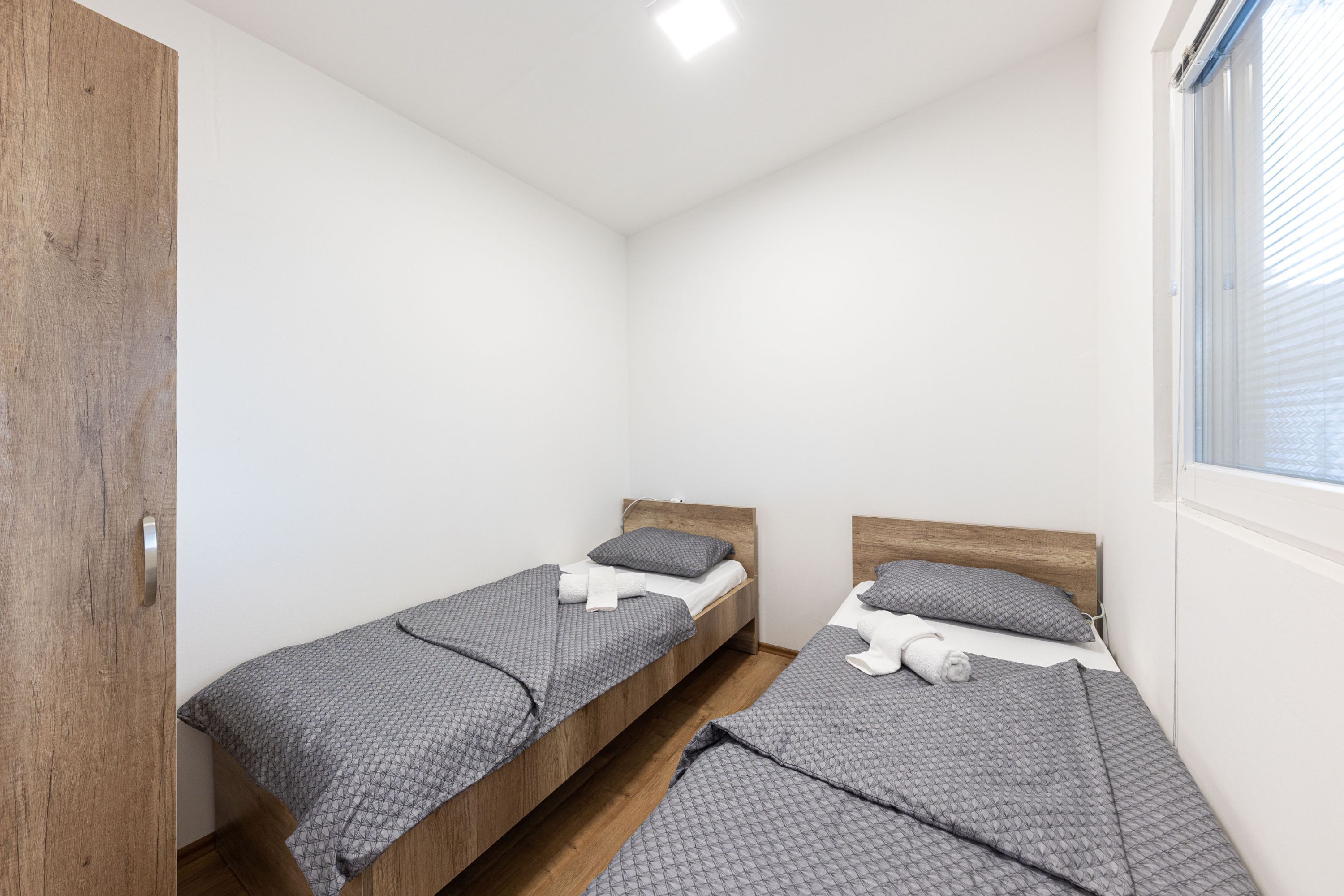 A small, clean room with two single beds, wooden headboards, and a window with blinds.