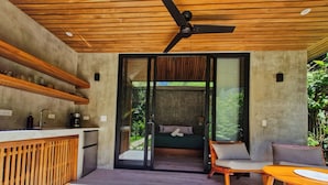 A modern kitchen with a wooden ceiling, a built-in fridge, and a coffee machine. There is a sliding glass door leading to a bedroom with a bed and a chair. - Serenity Sand Bungalow (Cahuita)