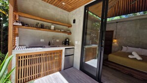 A compact, modern kitchen with wooden shelves, a sink, and a coffee maker. Adjacent is a glass door leading to a sleeping area with a bed and pillows. - Serenity Sand Bungalow (Cahuita)