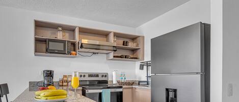 Una cocina moderna con un refrigerador de acero inoxidable, horno empotrado, microondas y una cafetera. Hay estantes de madera sobre la encimera y una encimera de granito.