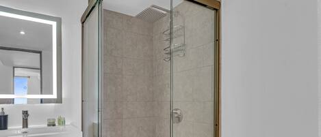 Un baño moderno con cabina de ducha de vidrio, un espejo grande y encimera blanca con artículos de aseo.