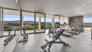 A modern gym with various exercise equipment, large windows offering a city view. - Cozy, Modern, Upscale APT W/Views, Pool, Gym, A/C (San Salvador)