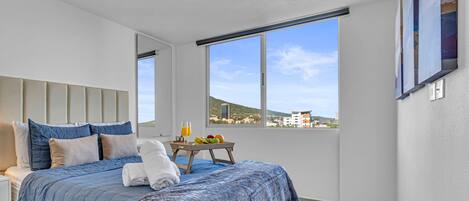 A modern hotel room with a large bed, a bedside table with a fruit bowl, and a view of the city through large windows.