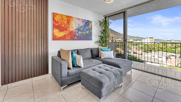 A modern living room with a grey sectional sofa, a large abstract painting, and a view of the city through a balcony. - Cozy, Modern, Upscale APT W/Views, Pool, Gym, A/C (San Salvador)