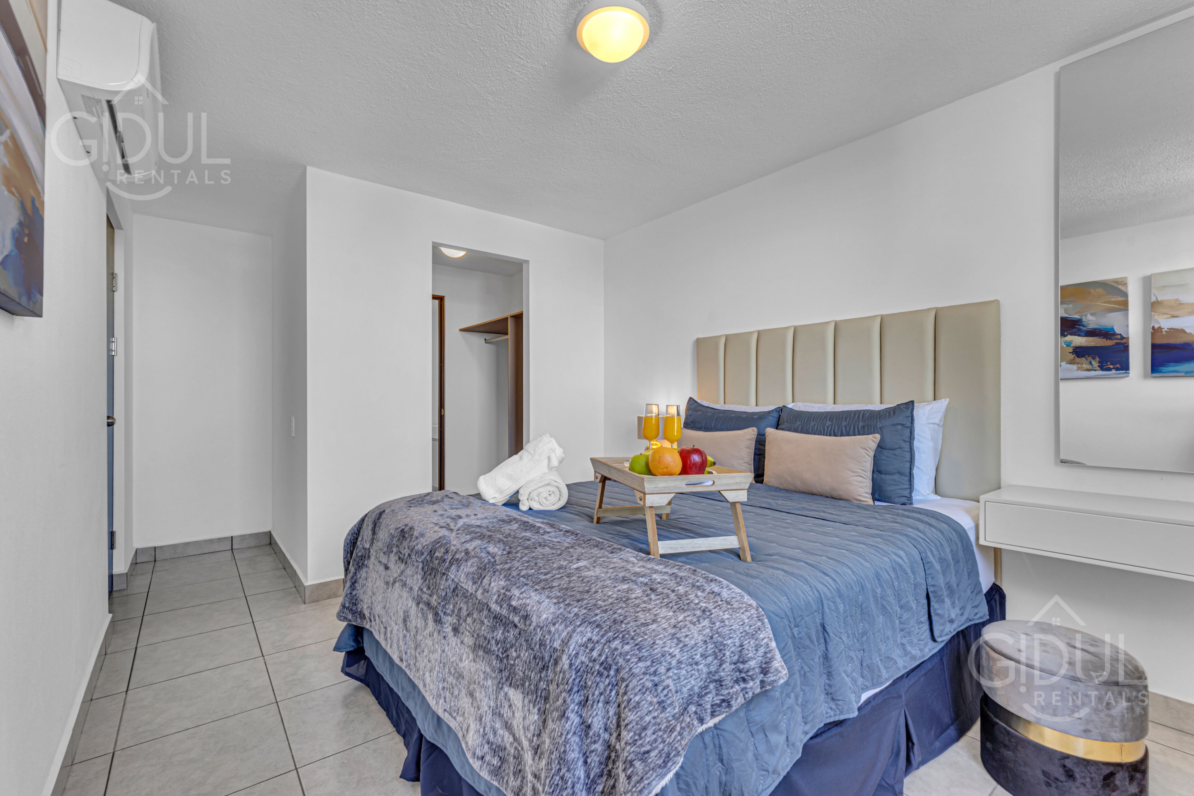 A bedroom with a bed, a bedside table with fruit and drinks, a towel, and a headboard.