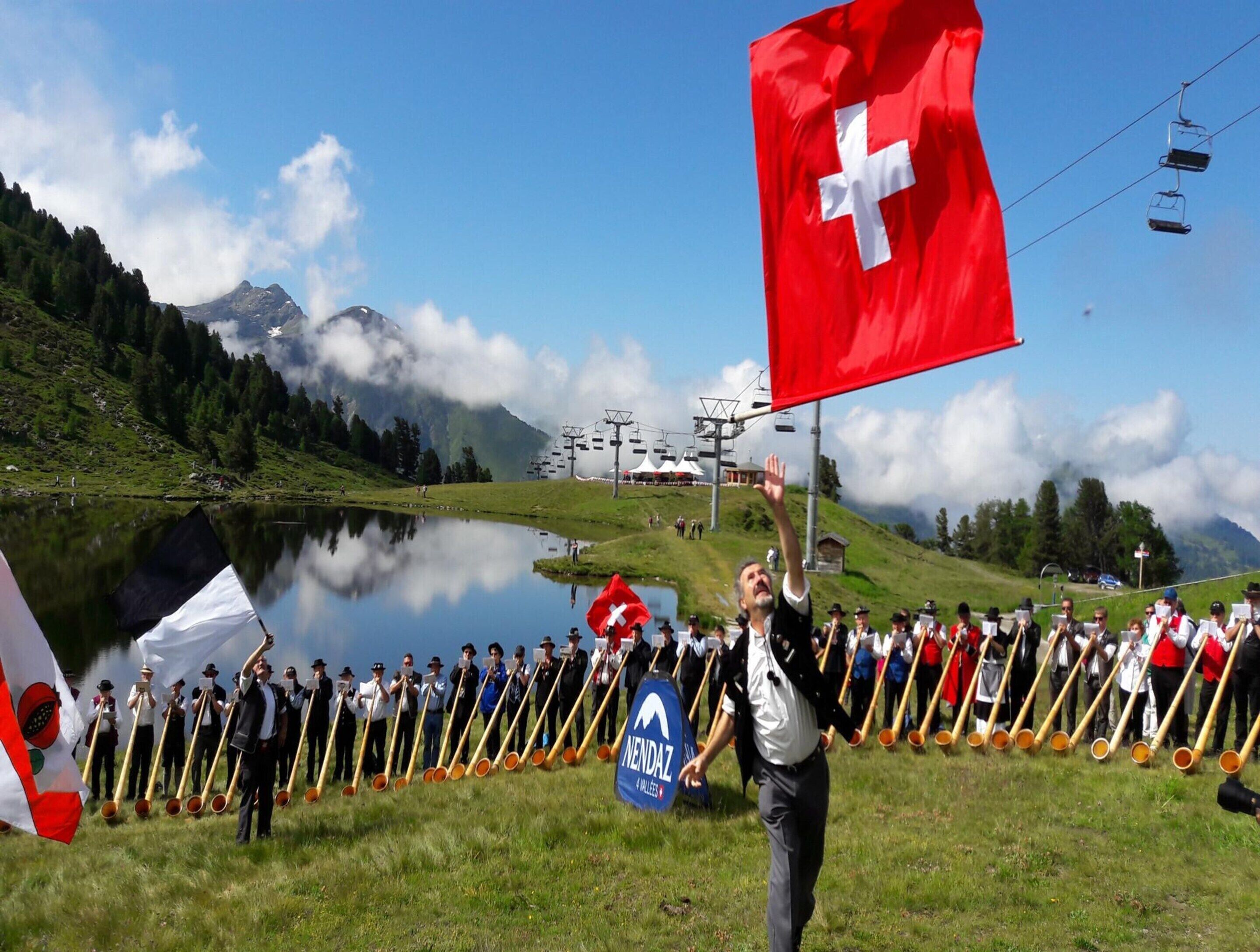 A group of people in traditional attire with long wooden sticks, standing in a line on a grassy field with a lake and mountains in the background.