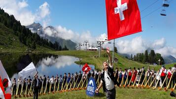 A group of people in traditional attire with long wooden sticks, standing in a line on a grassy field with a lake and mountains in the background.