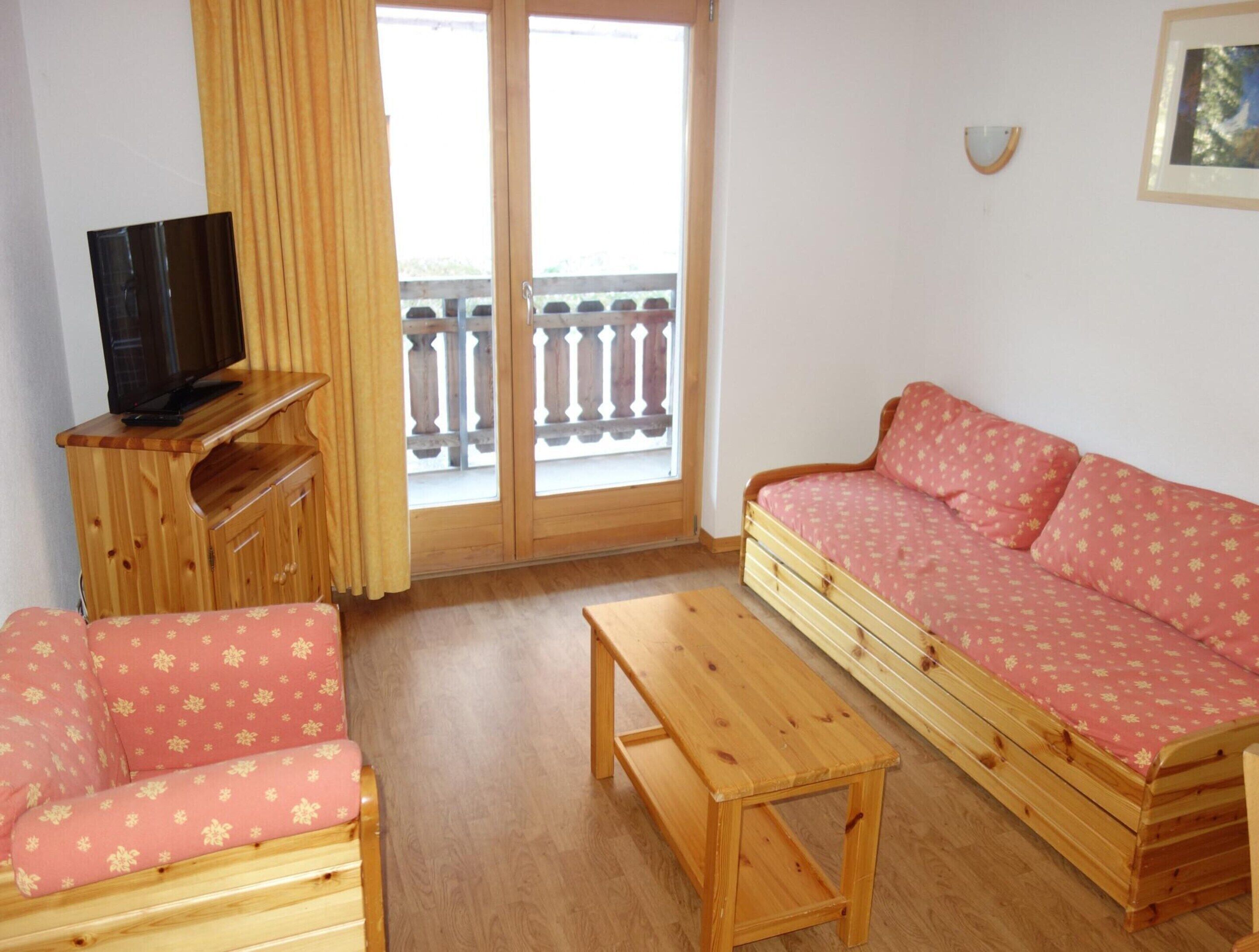 A room with a wooden sofa, a wooden table, a television on a wooden stand, and a balcony with a railing.