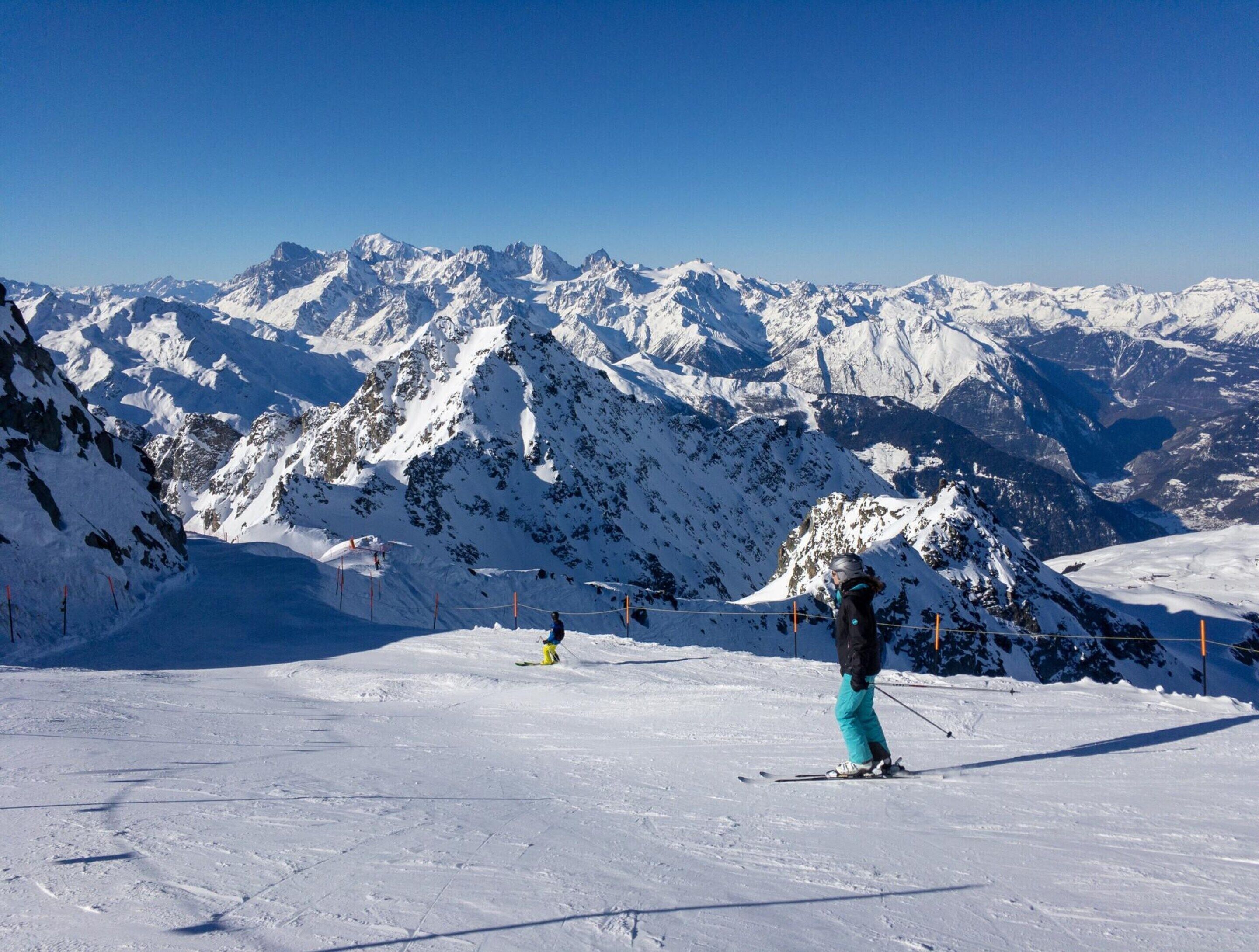 Une piste de ski enneigée où l’on voit une personne skier, entourée de montagnes recouvertes de neige sous un ciel dégagé.