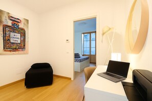 Una habitación moderna con un ordenador portátil sobre un escritorio, un reposapiés negro, una lámpara de pared y una puerta que da a otra habitación.