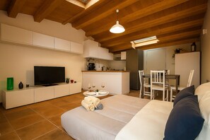 Un ambiente compatto con divano, televisore, tavolo da pranzo con sedie, angolo cottura e soffitto con travi in legno.