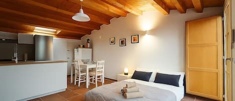A bedroom with a bed, a dining table, chairs, a kitchen area, and a wooden ceiling.