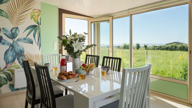 A dining area with a table set for four, a vase of flowers, and a view of a green field through large windows. - Magic Mountain breathtaking view of Mt Stmichel Bay (Dragey-Ronthon)