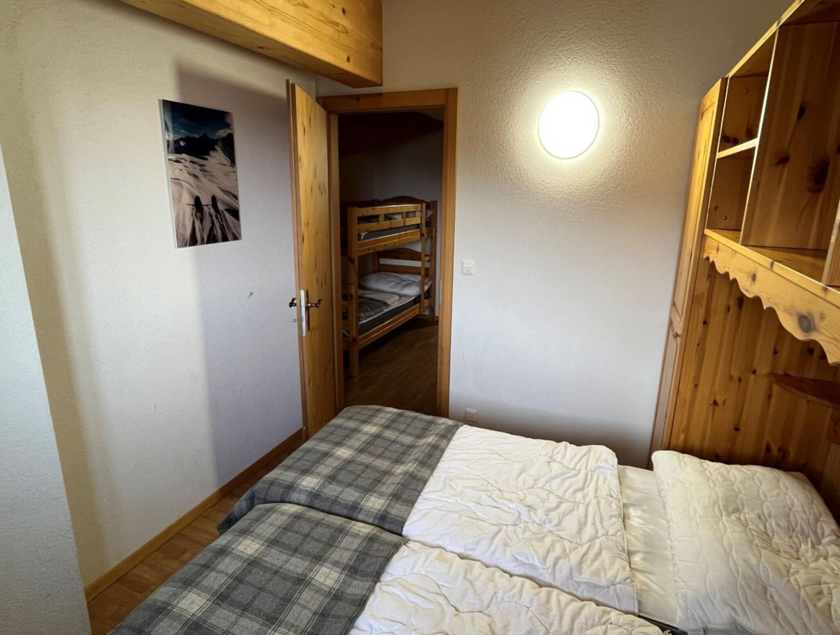 A bedroom with a bunk bed, a bed with plaid bedding, a wooden ceiling, and a wall-mounted light fixture.