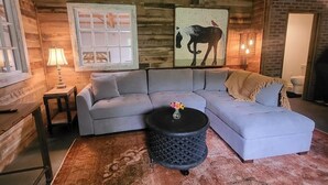 A cozy living room with a light gray sectional sofa, a round coffee table with a vase of flowers, and a rustic wooden wall with a horse painting. - Cozy loft inside a vintage store on an animal rescue farm in Westville, IN (Westville)
