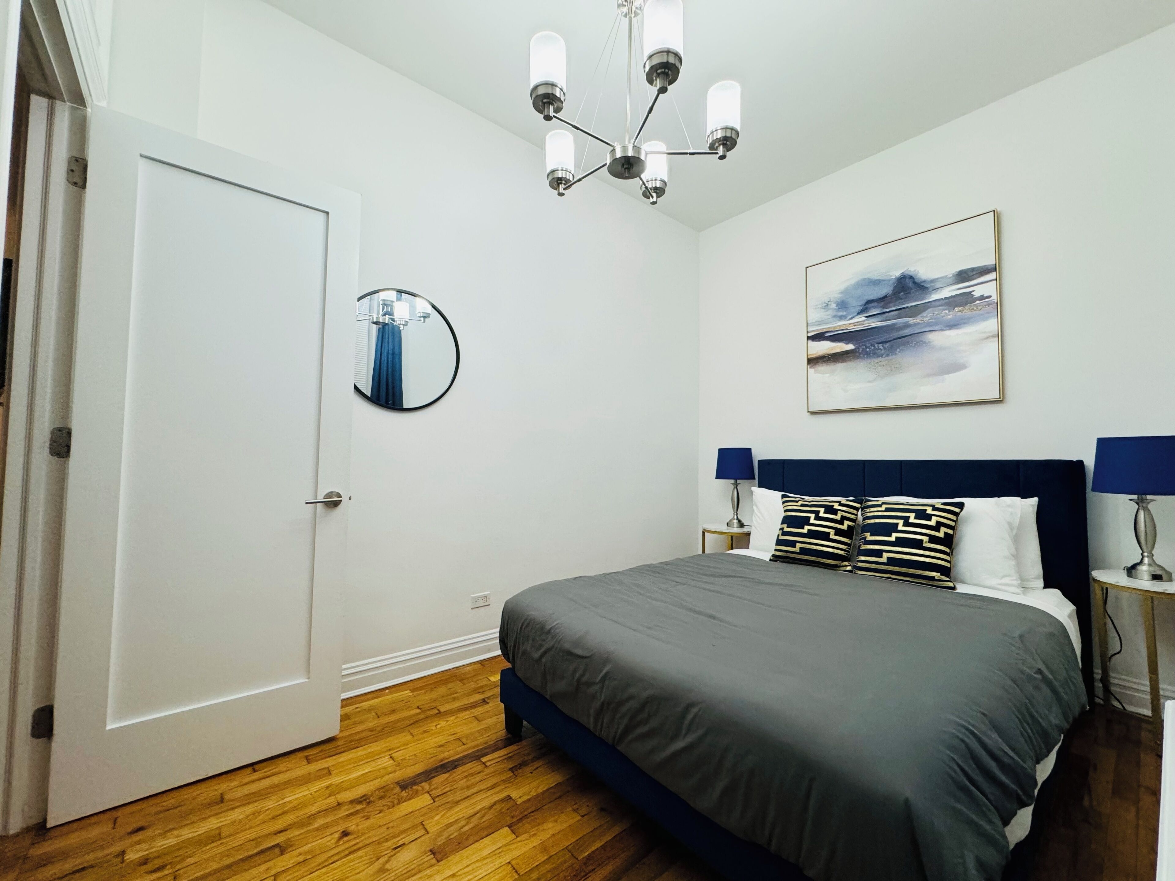 A bedroom with a bed, a round mirror, a chandelier, a painting, and a door.
